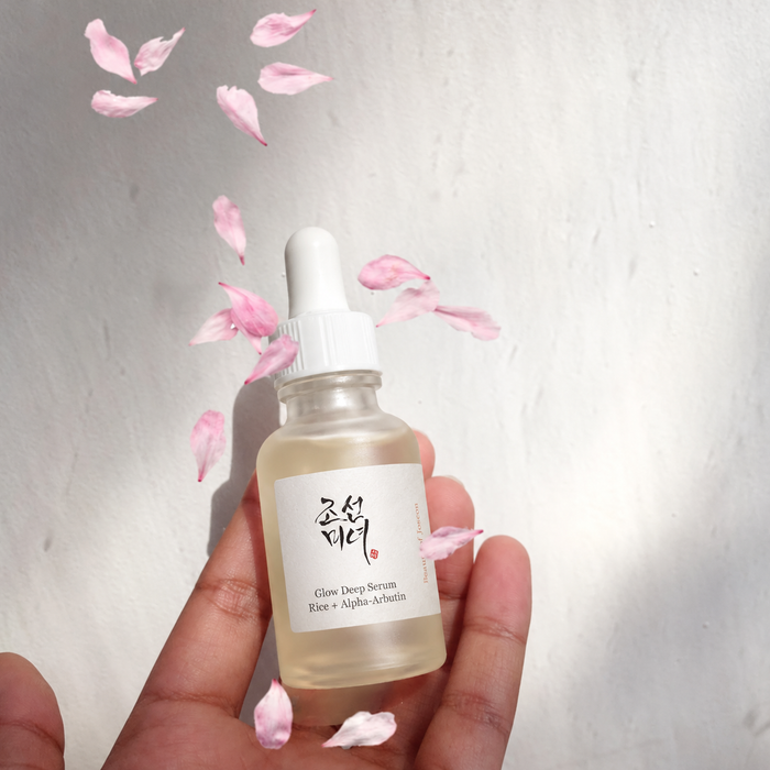 Beauty of Joseon Glow Deep Serum with Rice + Alpha-Arbutin held in a hand, surrounded by soft pink flower petals against a bright white background.